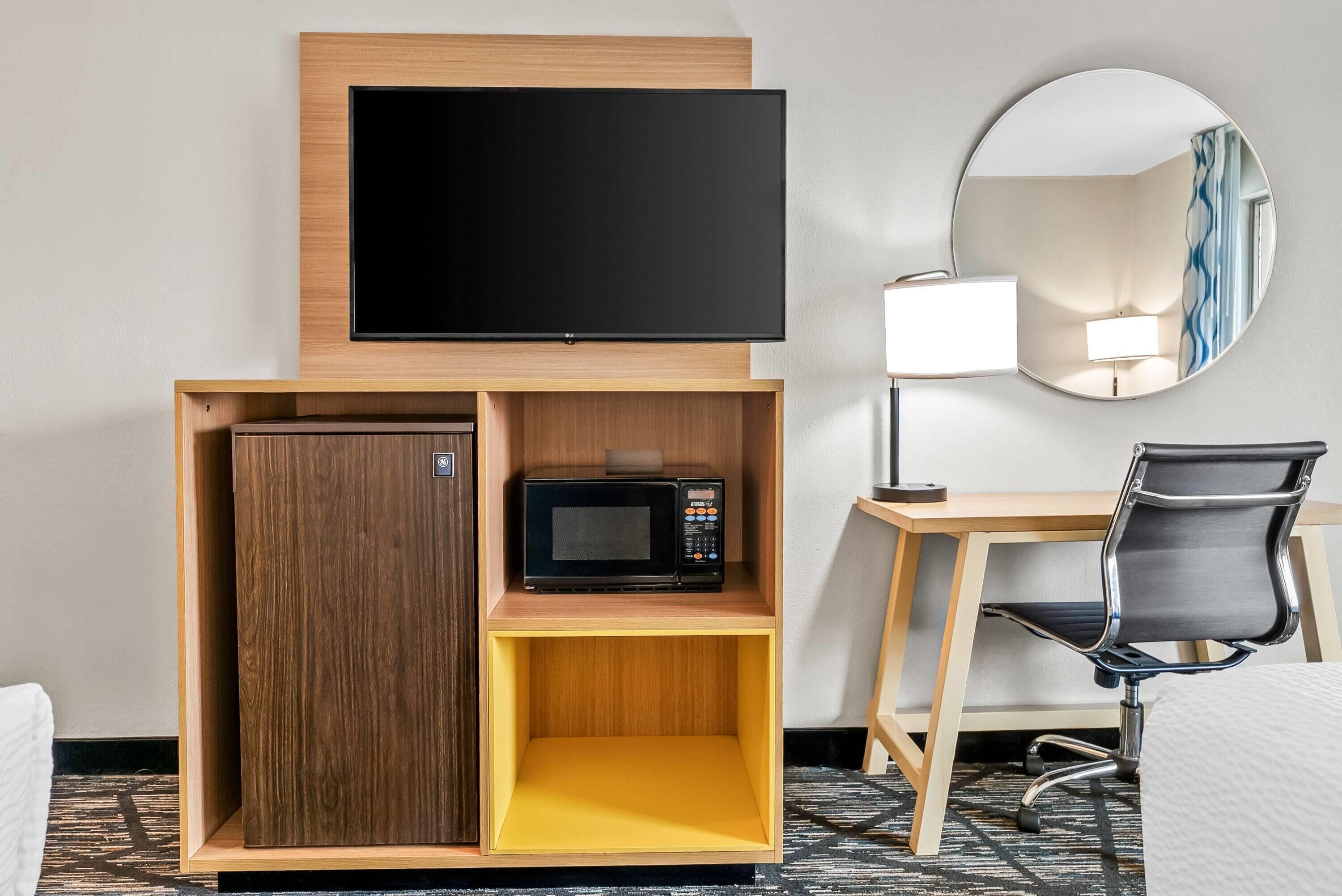 A hotel room with a flat-screen TV, a microwave, a small cabinet, a desk, a chair, and a round mirror.