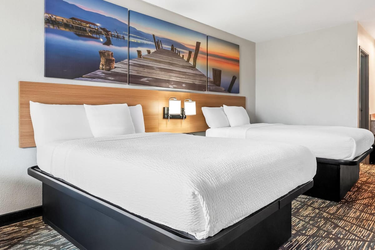 Ein Hotelzimmer mit zwei Betten, einem Holzkopfende, einem Nachttisch mit Lampe und einem großen Gemälde eines Piers im Sonnenuntergang.