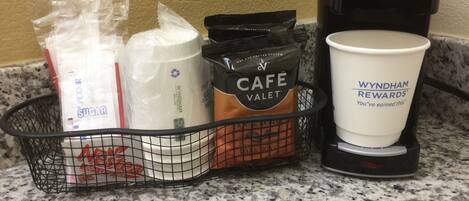 A coffee maker, a cup, and a basket with coffee bags and sugar packets.