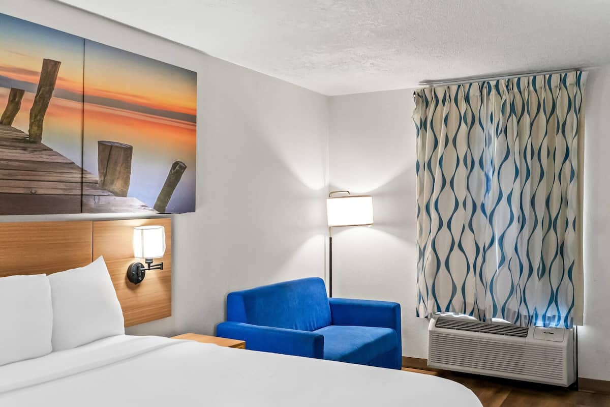 Ein Hotelzimmer mit einem Bett, einem blauen Sessel, einer Wandlampe, einem Sonnenuntergangsgemälde und einem gemusterten Vorhang.
