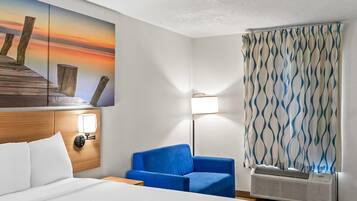Habitación de hotel con cama, un sillón azul, una lámpara de pared, un cuadro de un atardecer y una cortina estampada.