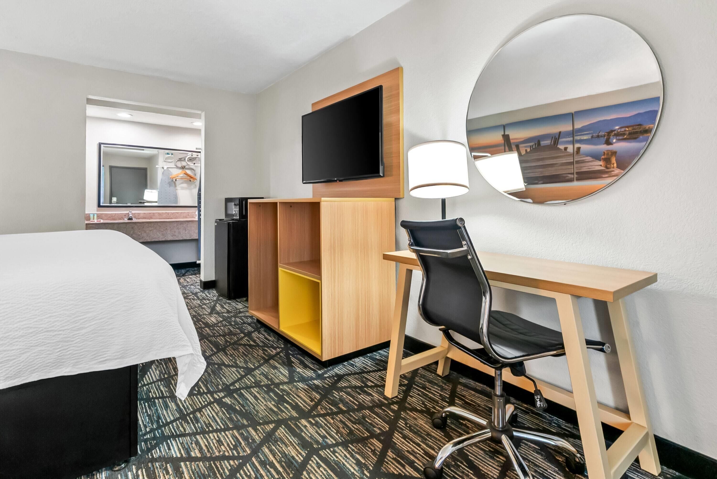 A hotel room with a bed, a desk with a chair, a TV, a mirror, and a painting on the wall.