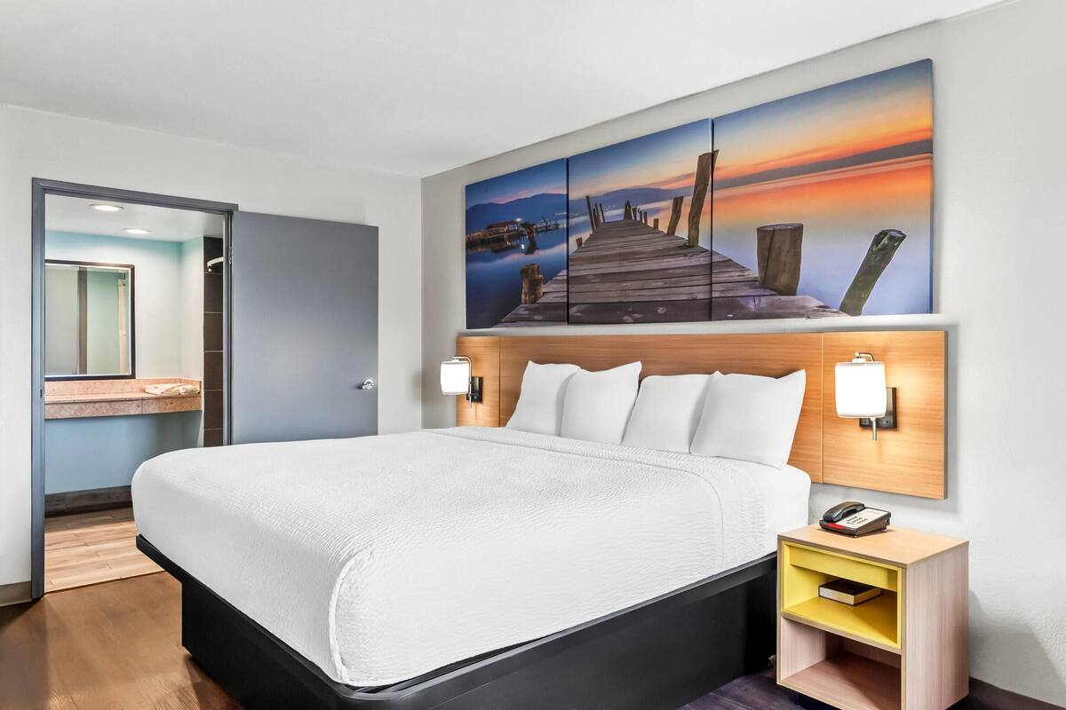 Ein Hotelzimmer mit einem großen Bett, einem Nachttisch mit Lampe, einem Gemälde eines Piers und einem Holz-Kopfteil.