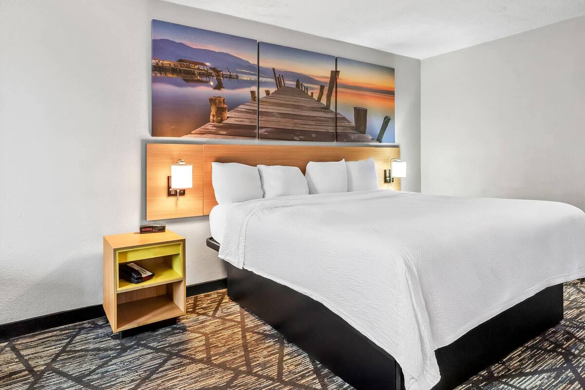 Ein Hotelzimmer mit einem großen Bett, einem hölzernen Kopfteil, zwei Nachttischlampen, einem kleinen Nachttisch und einem großen Wandbild, das einen Pier bei Sonnenuntergang zeigt.