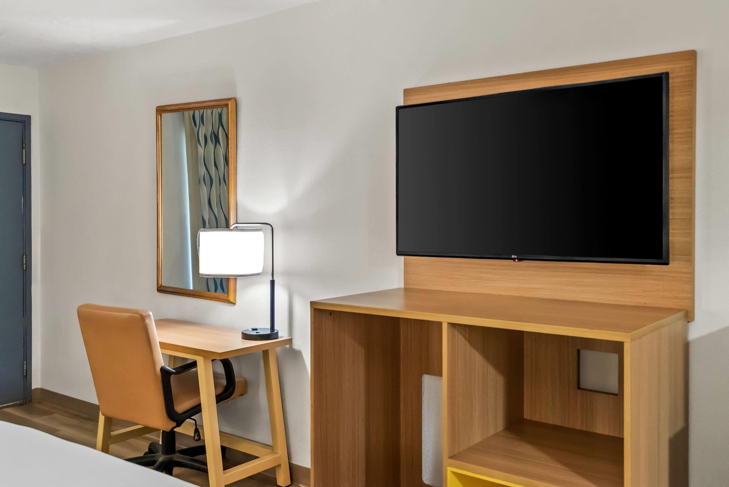 A hotel room with a wooden desk, a chair, a flat-screen TV mounted on the wall, and a mirror with a curtain.