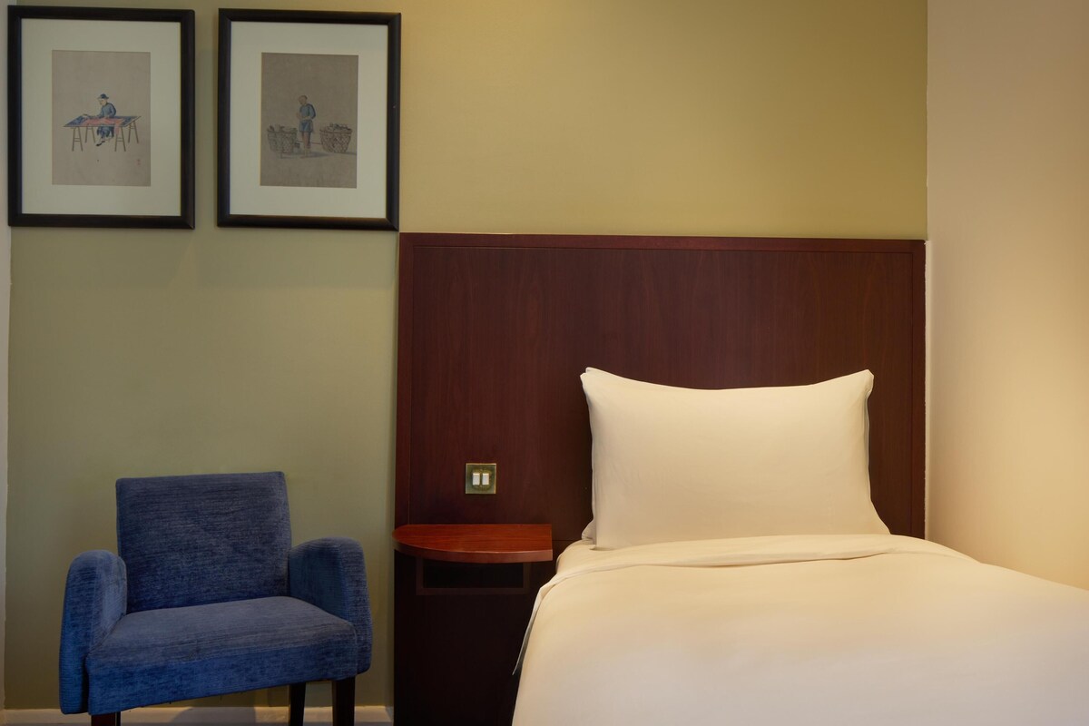 A hotel room with a bed, a blue armchair, a wooden nightstand, and two framed pictures on the wall.