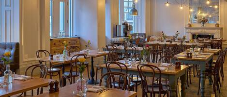 A spacious dining area with multiple wooden tables and chairs, large windows, and hanging pendant lights.