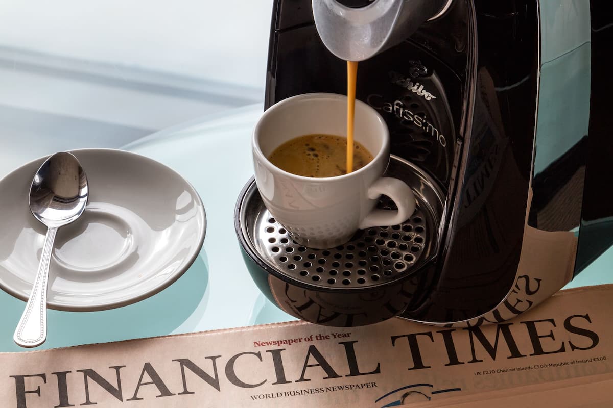 A coffee machine pouring coffee into a cup, a spoon, and saucer on a table with newspapers.