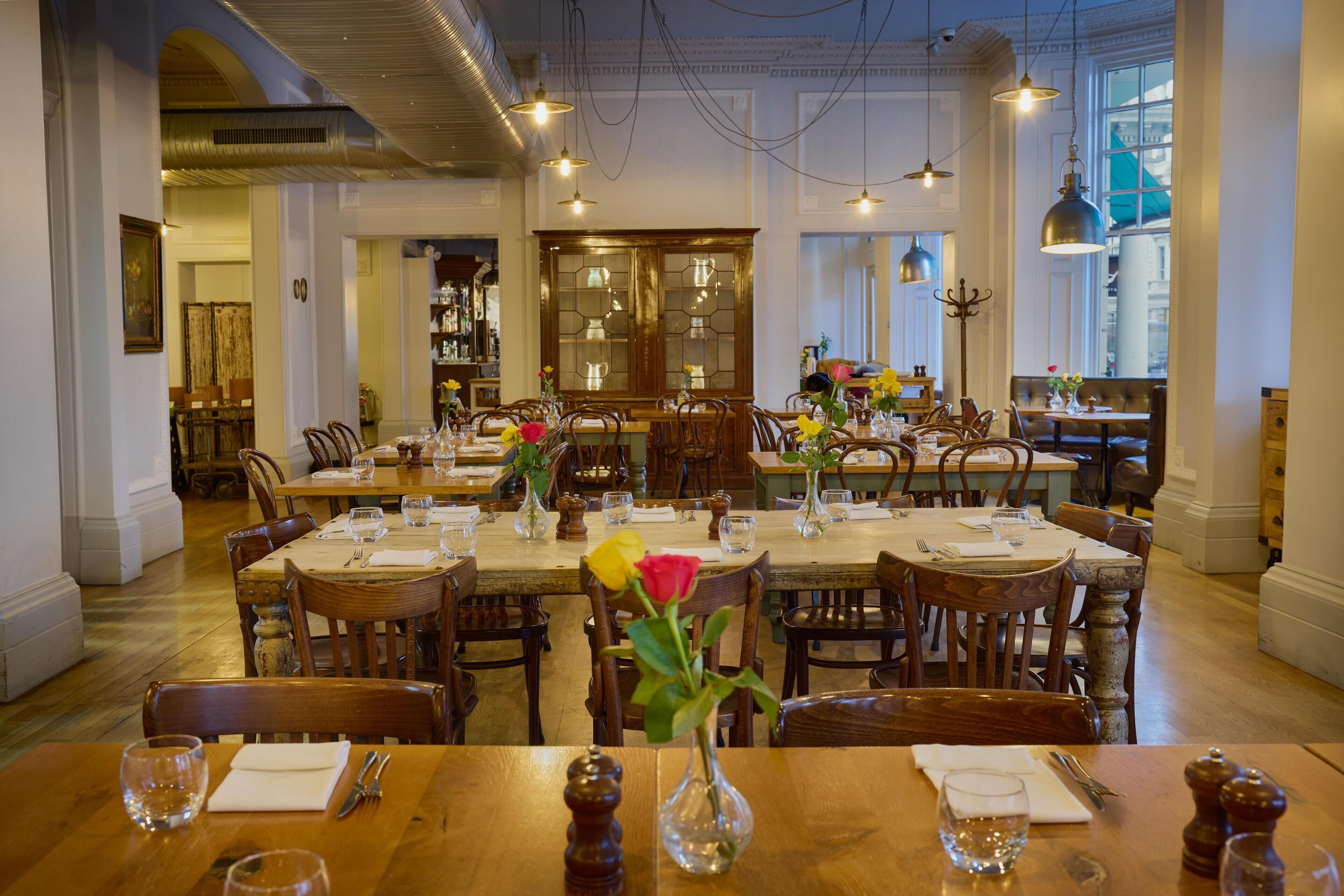 A dining area with wooden tables and chairs, a central dining table with a vase of flowers, and a large cabinet in the background.