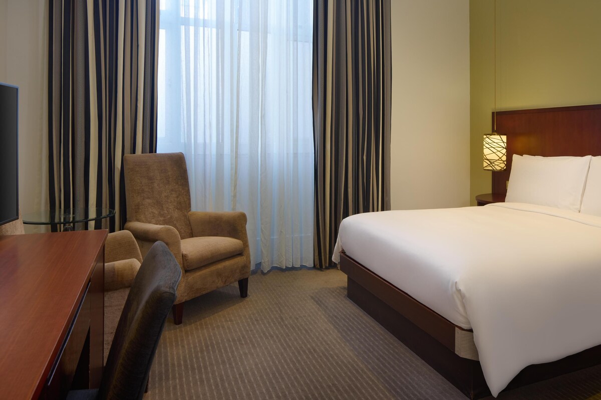 A hotel room with a bed, a chair, a desk, and a lamp.