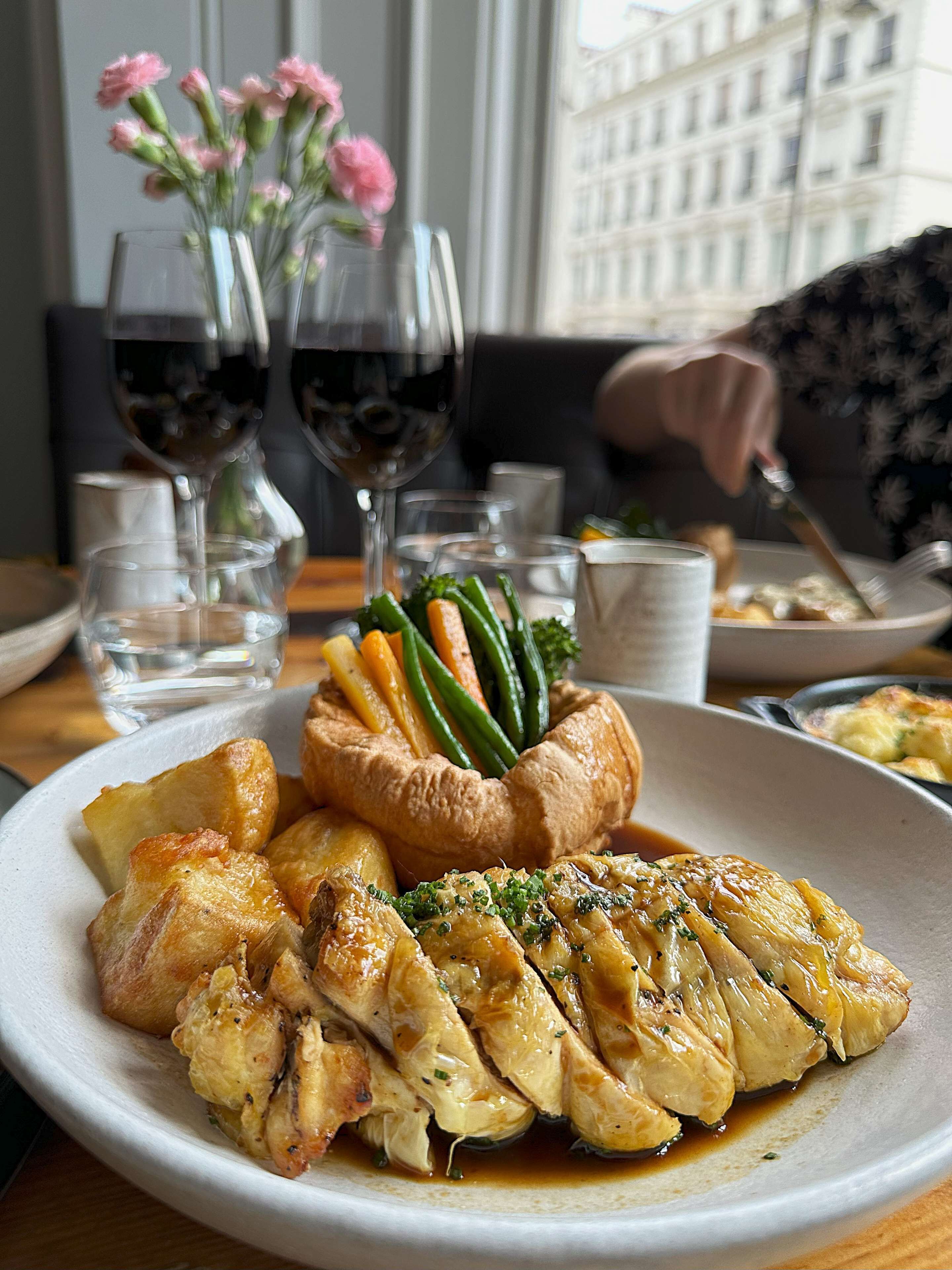 A plate of food with roasted chicken, Yorkshire pudding, and vegetables, accompanied by wine glasses and a vase of flowers.