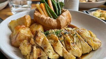 A plate of food with roasted chicken, Yorkshire pudding, and vegetables, accompanied by wine glasses and a vase of flowers.