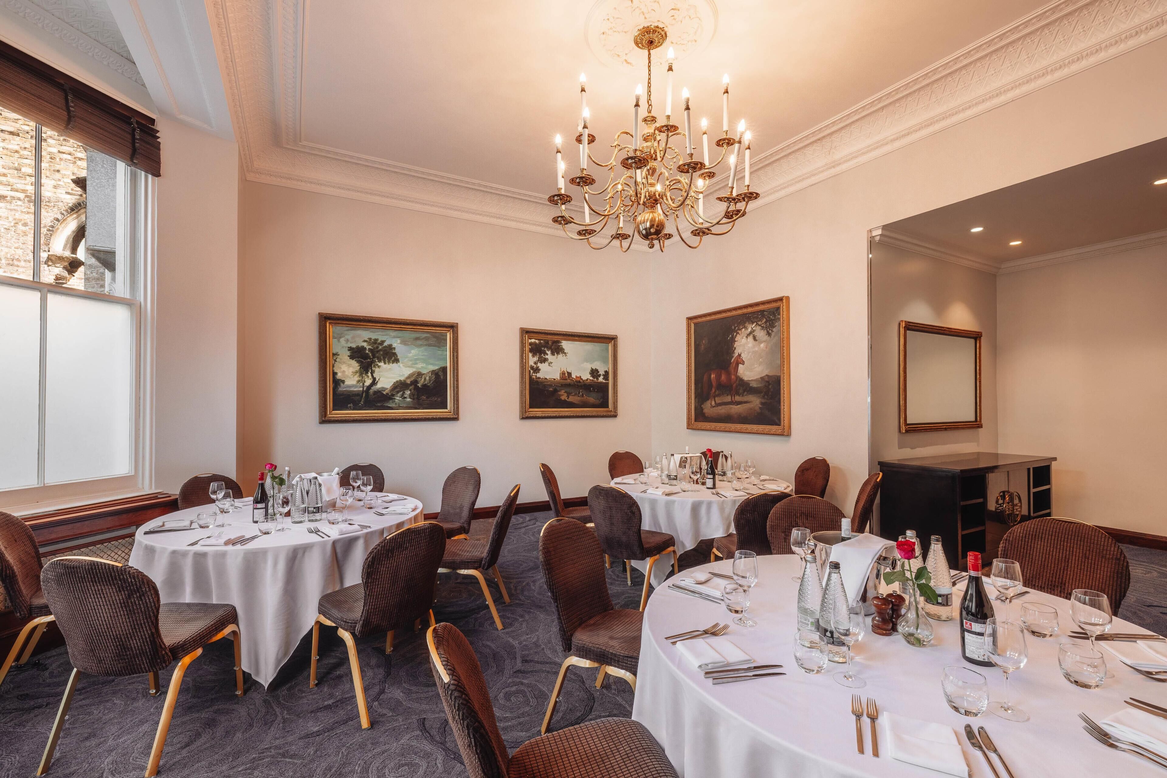 A dining area with round tables, chairs, chandeliers, framed paintings, and a carpeted floor.