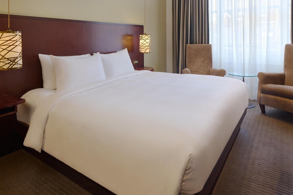 A hotel room with a large bed, two bedside lamps, a chair, and a small table.