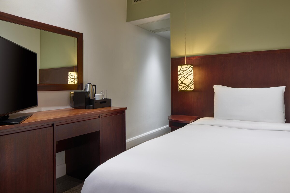 A hotel room with a wooden desk, a television, a bedside table with a lamp, and a bed with white bedding.