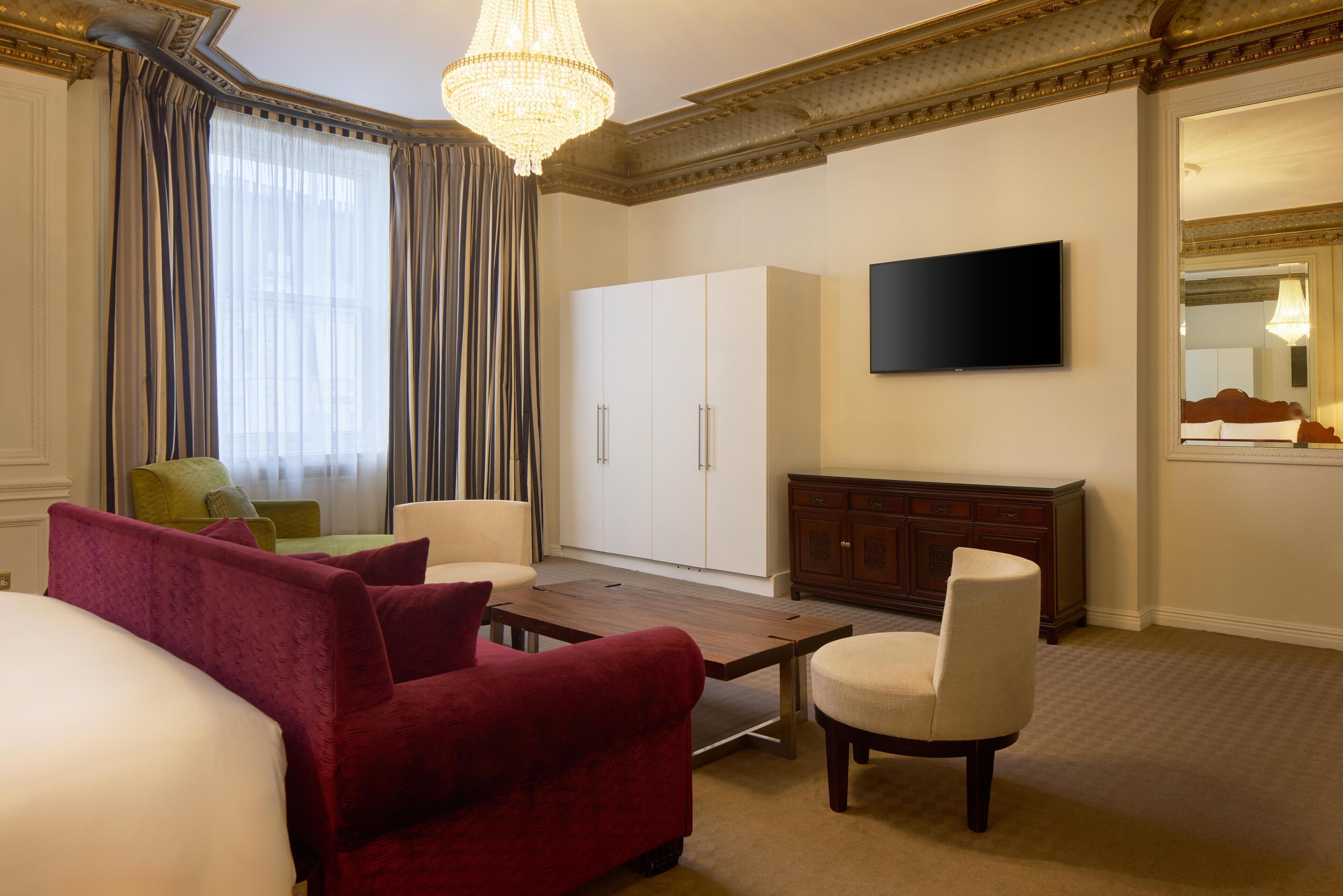 A hotel room with a bed, a red armchair, a white armchair, a wooden coffee table, a television, a wardrobe, and a large window with curtains.