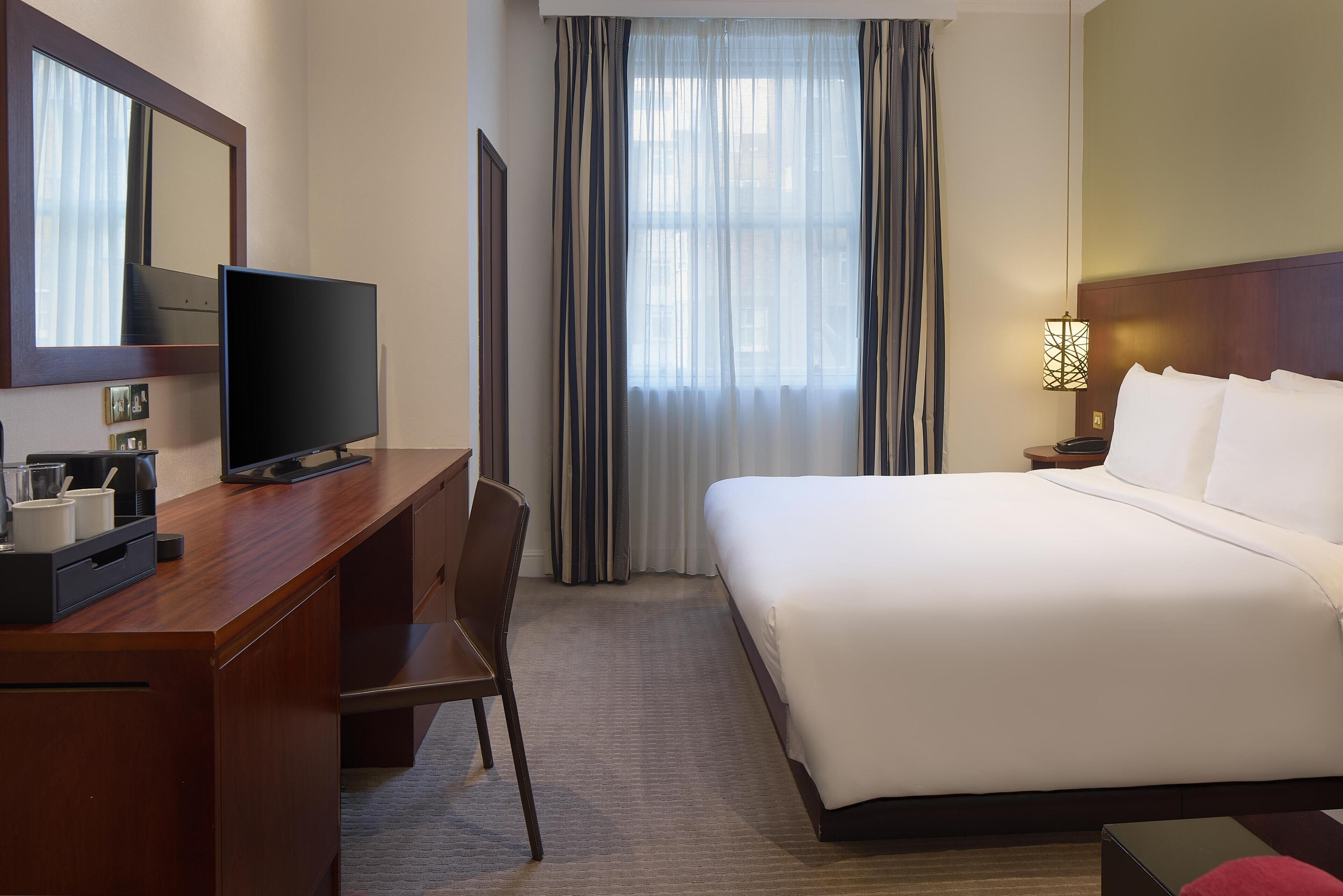 Een hotelkamer met een houten bureau, een televisie, een bed met witte lakens, een nachtkastje met een lamp en een raam met gordijnen.