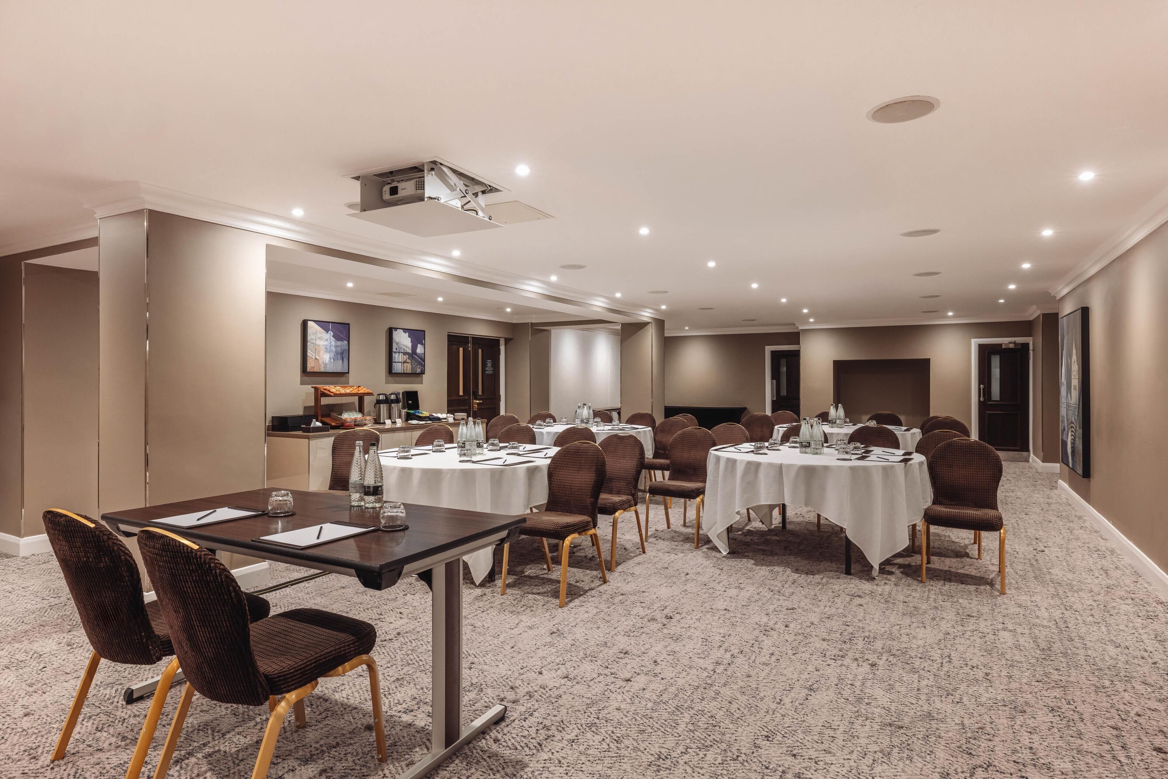 A conference room with round tables, chairs, a projector, and a kitchenette area.