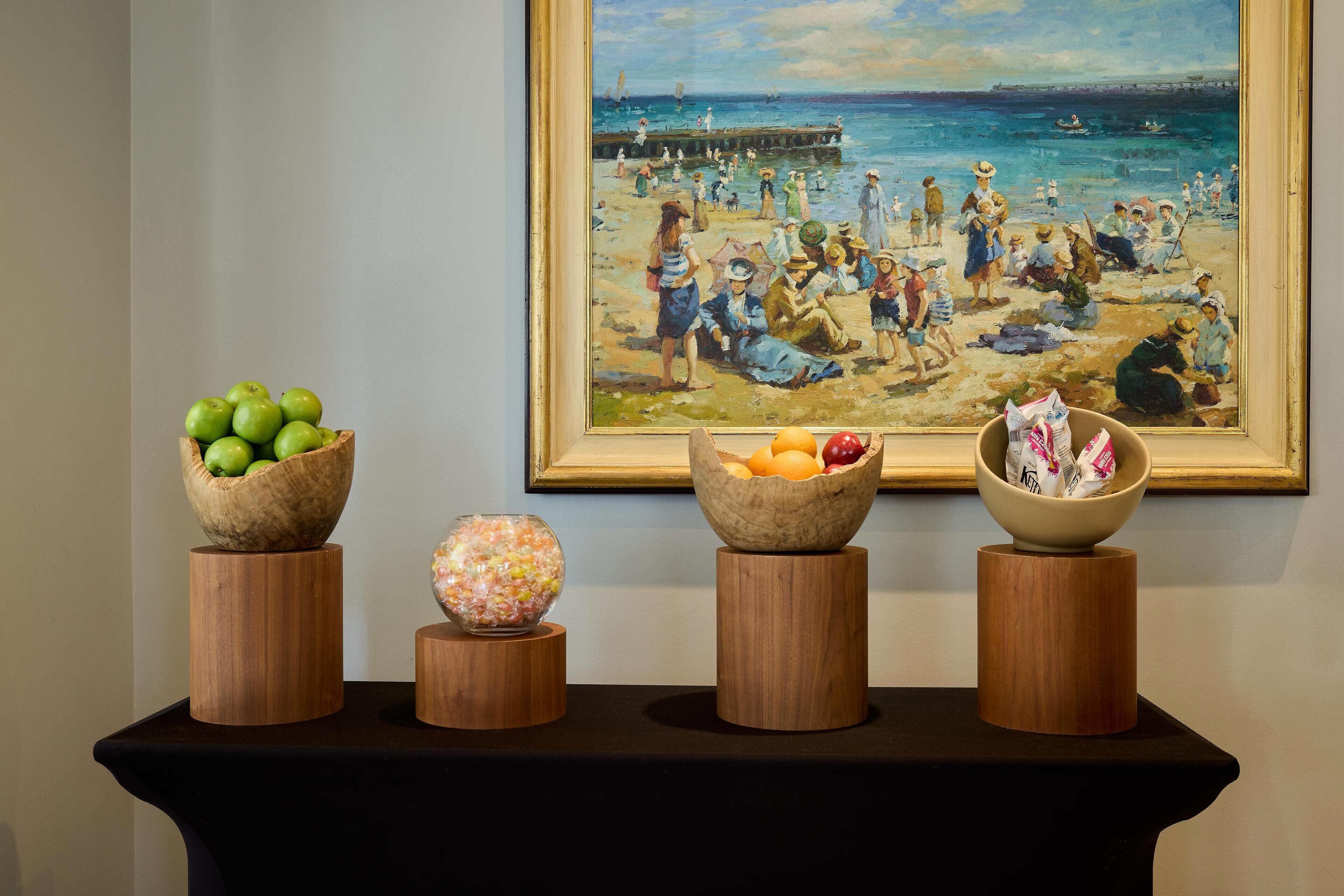 A painting of a beach scene with people and boats, and three wooden stands with bowls of fruit and a decorative glass.