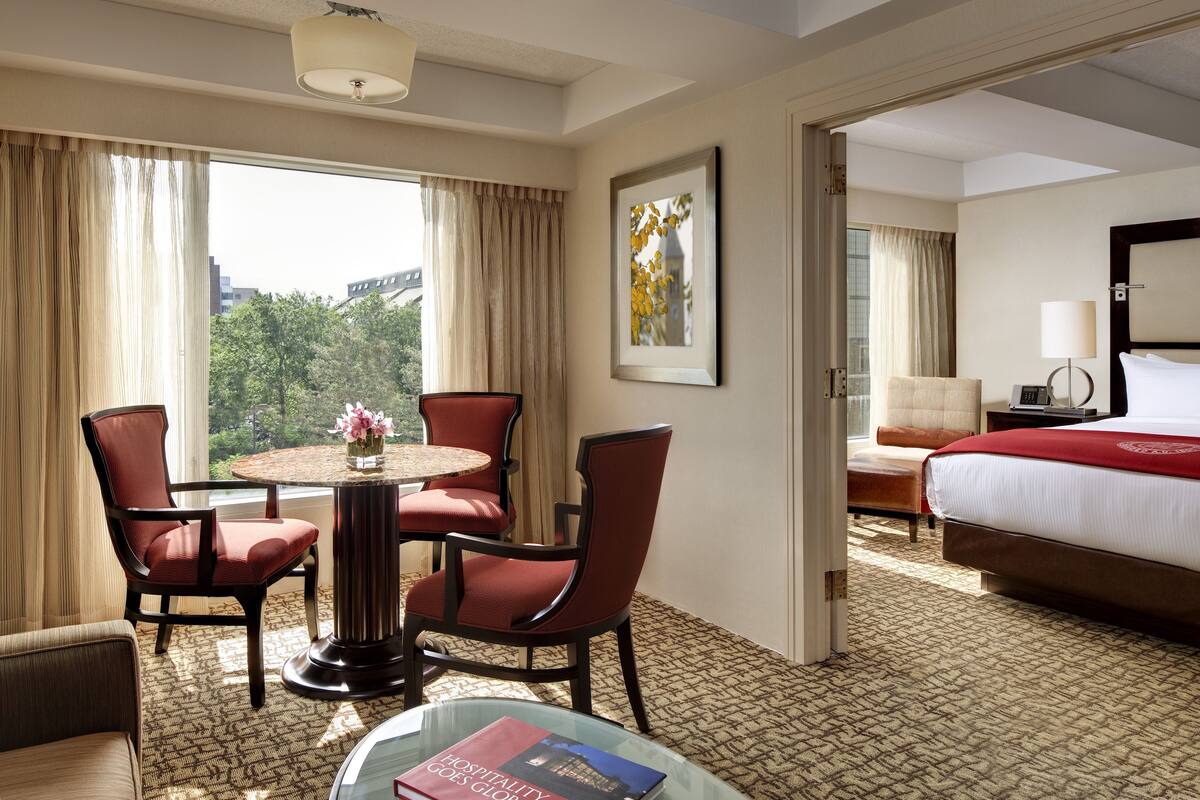 A hotel room with a bed, a round table with chairs, a sofa, a bedside table with a lamp, and a framed picture on the wall.