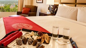 Een hotelkamer met een bed, een dienblad met chocolade en een welkomstbord, twee glazen water en uitzicht op een stad door het raam.