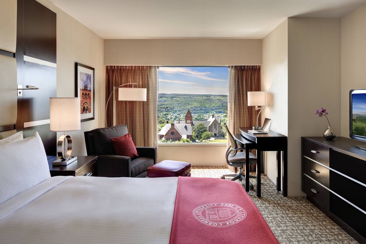 A hotel room with a large bed, a desk, a chair, and a view of a town with a church.