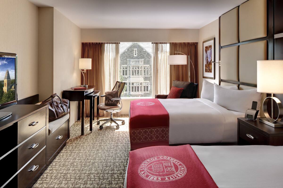 A hotel room with a large bed, a desk, a chair, and a view of a building through the window.