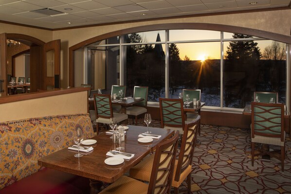 Un comedor con una mesa grande dispuesta para cuatro personas, rodeada de sillas con respaldo acolchado. Un amplio ventanal permite ver el atardecer y los árboles al exterior.