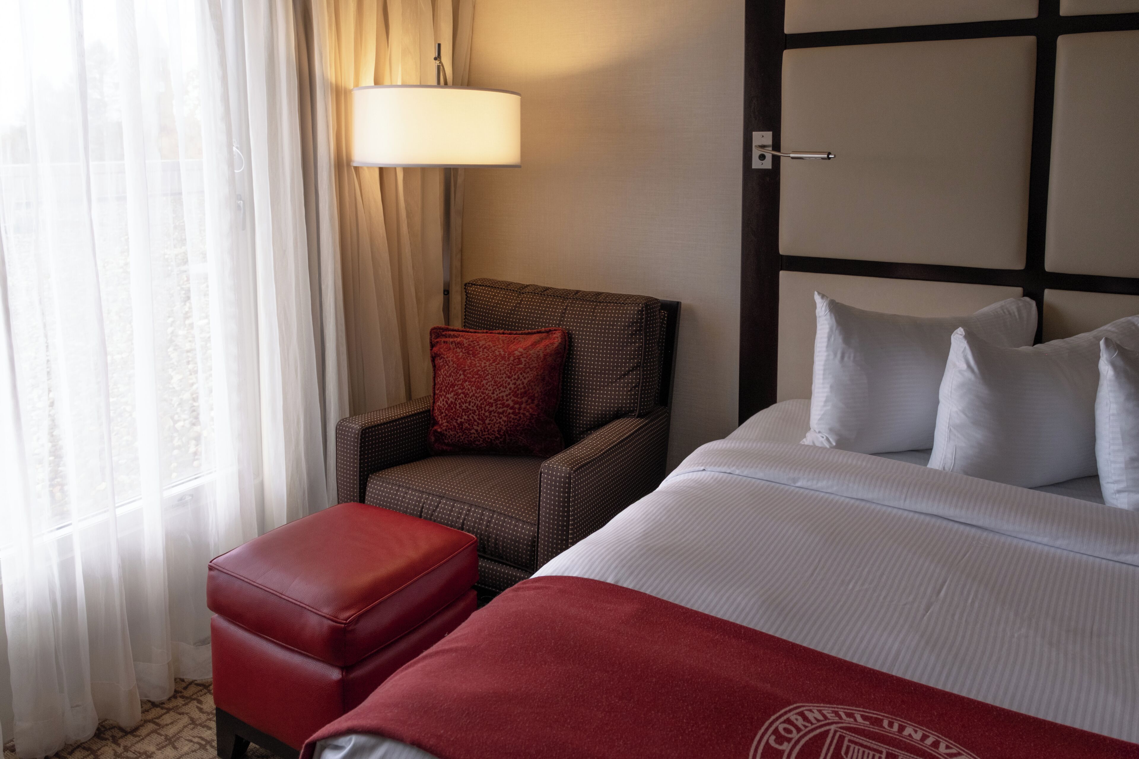 A hotel room with a bed, a chair, a red ottoman, a lamp, and a window with curtains.