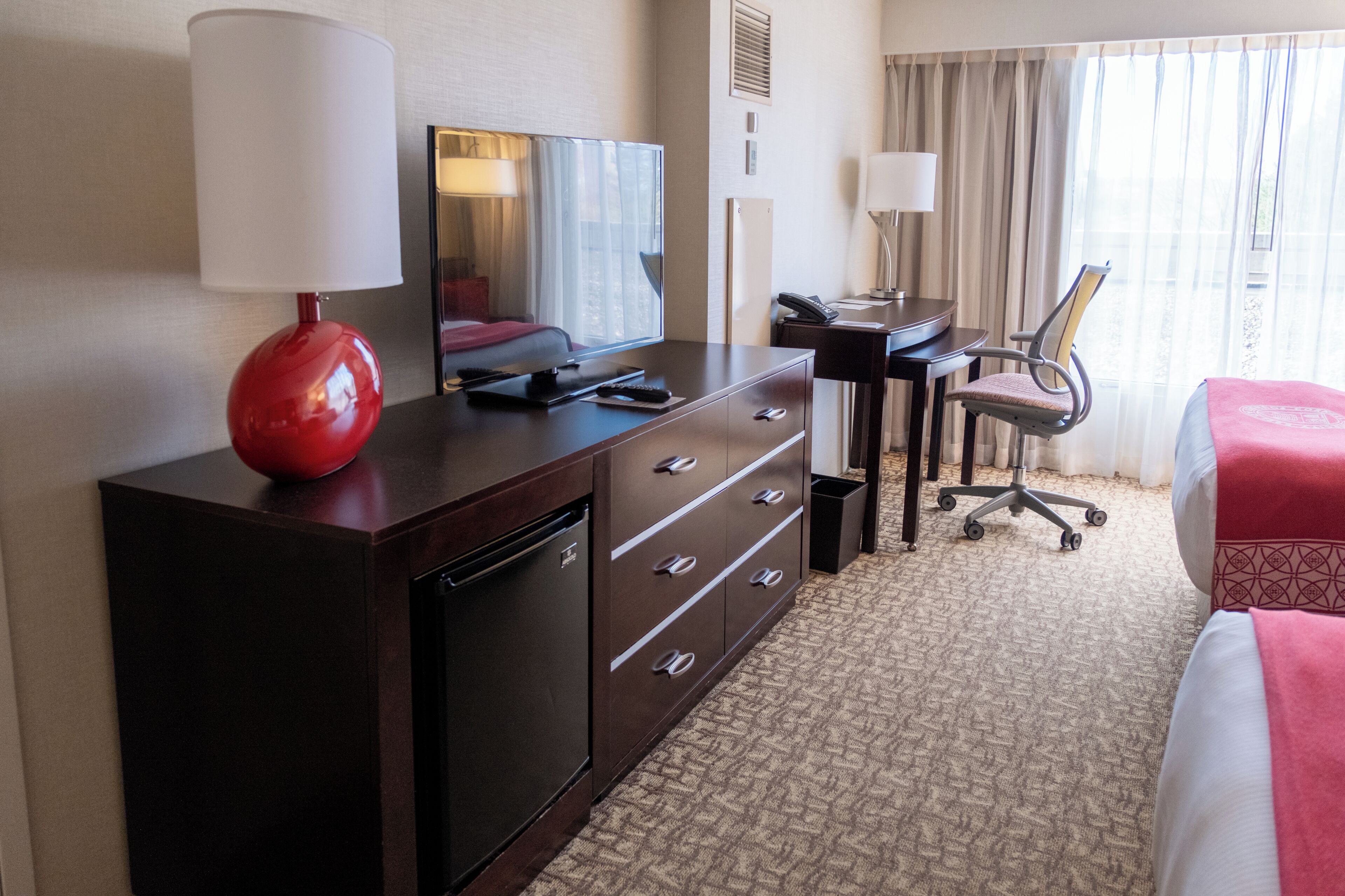 A hotel room with a bed, a desk with a chair, a TV, a lamp, and a dresser.