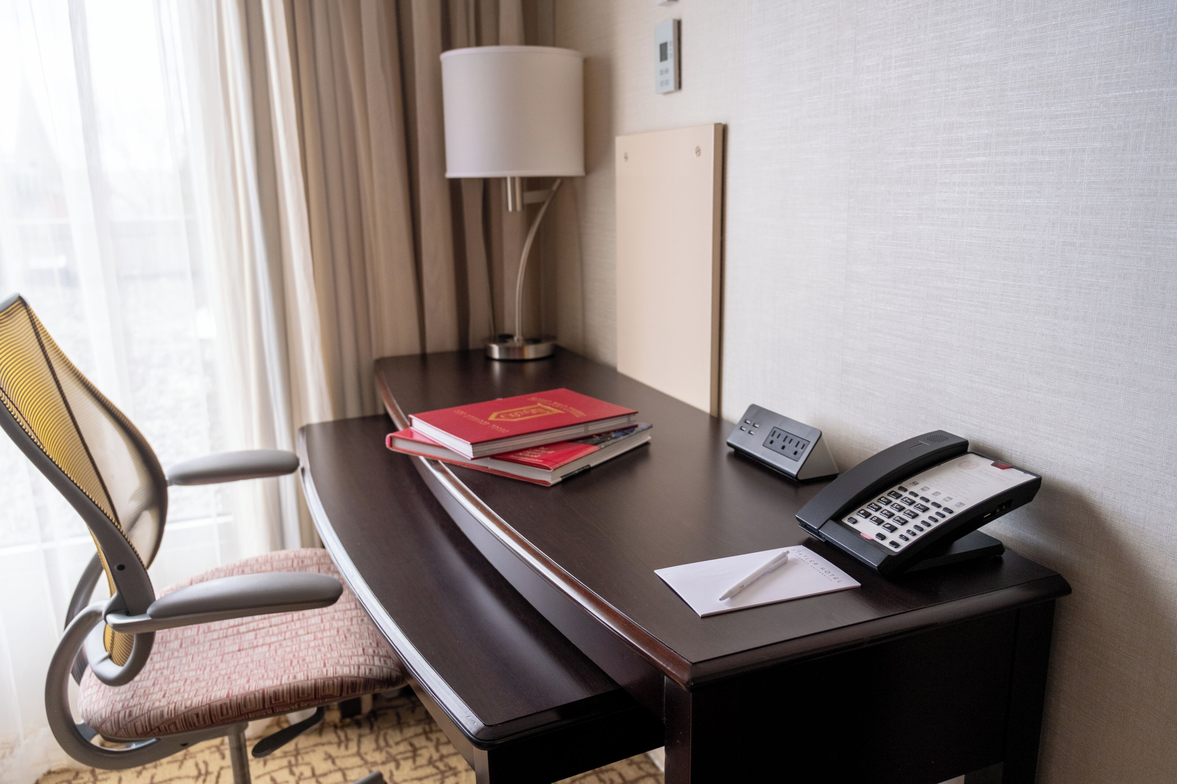 A hotel room with a desk, a telephone, a lamp, and a chair.