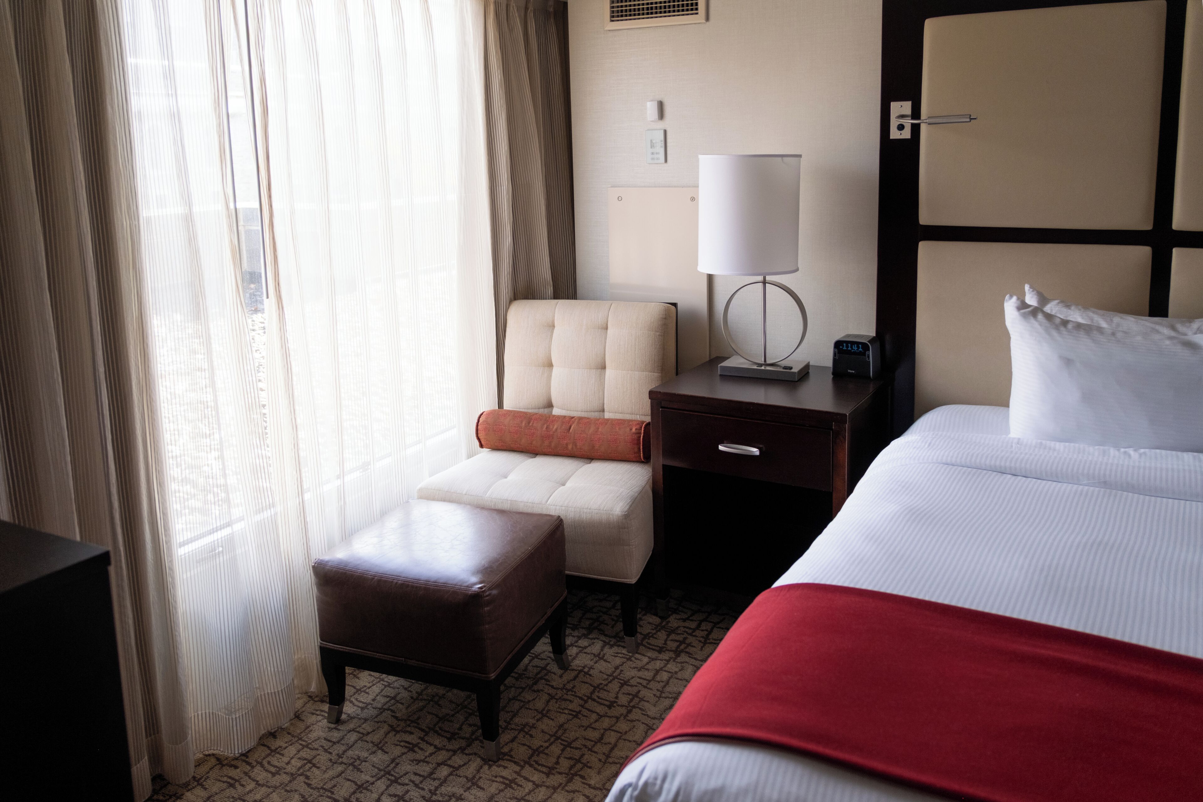 A hotel room with a bed, a chair, an ottoman, a nightstand, a lamp, and a window with curtains.