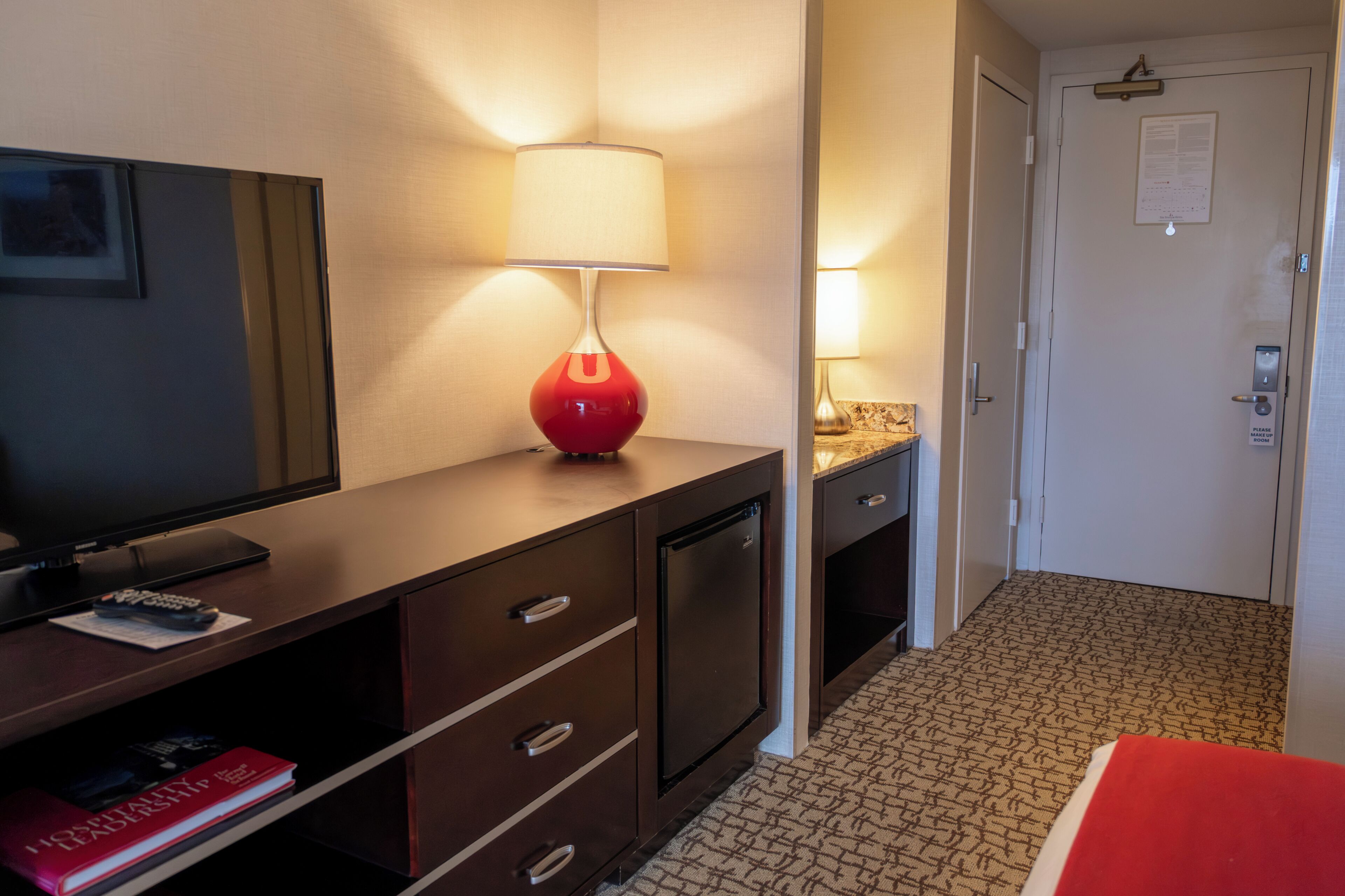 A hotel room with a TV, a red lamp, a minibar, a door with a lock, and a patterned carpet.