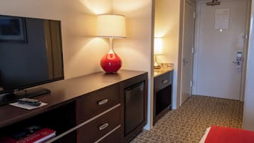A hotel room with a TV, a red lamp, a minibar, a door with a lock, and a patterned carpet.