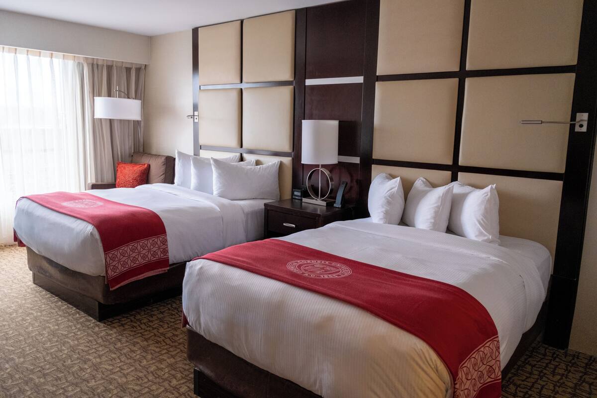 A hotel room with two beds, a large window with curtains, a nightstand with a lamp, and a wall panel design.