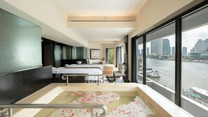 A hotel room with a large bathtub filled with rose petals, a bed with white linens, and a view of a river and cityscape through large windows. - Royal Orchid Sheraton Riverside Hotel Bangkok (Bangkok)