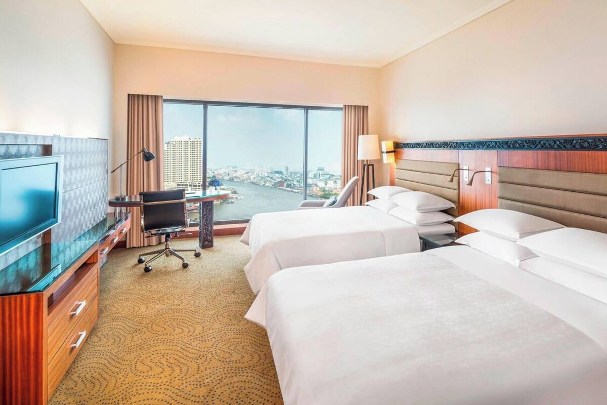 Habitación de hotel con dos camas, un escritorio, una silla, un televisor y un ventanal con vistas a la ciudad.