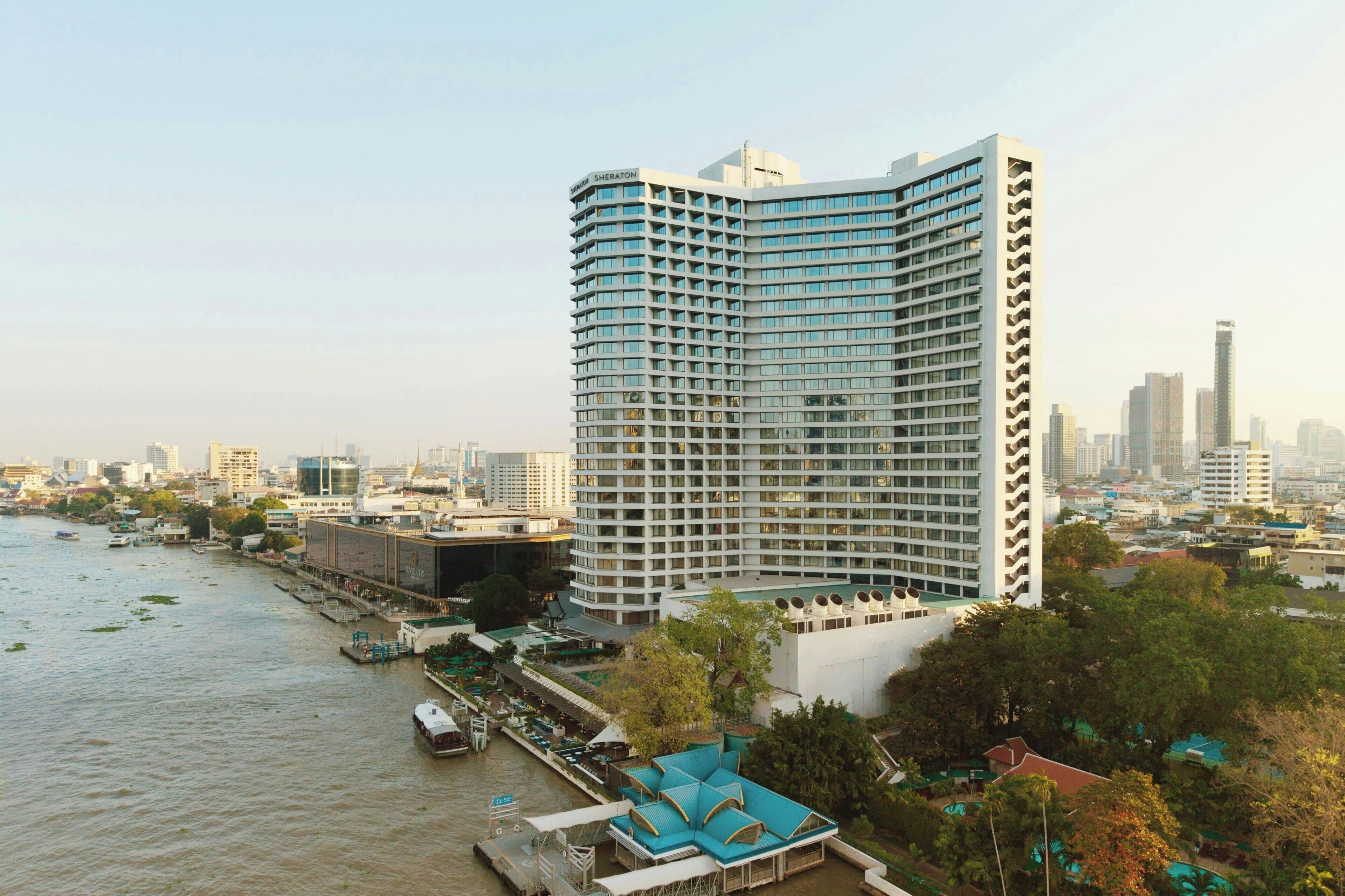 Un hotel moderno de altura con fachada curva, situado cerca de un río con un embarcadero y barcos.