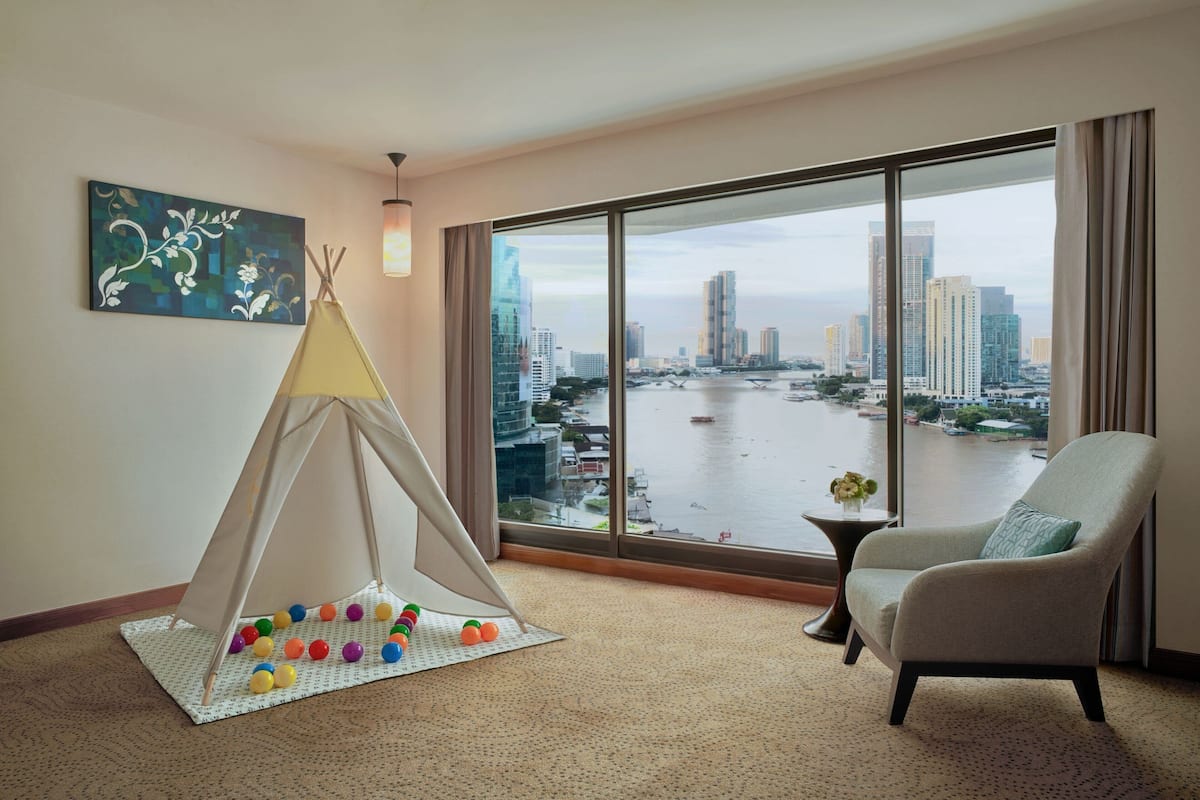 Una habitación con un ventanal grande que ofrece vistas de la ciudad, una silla, una mesita con un florero y una tienda tipi con pelotas de colores.