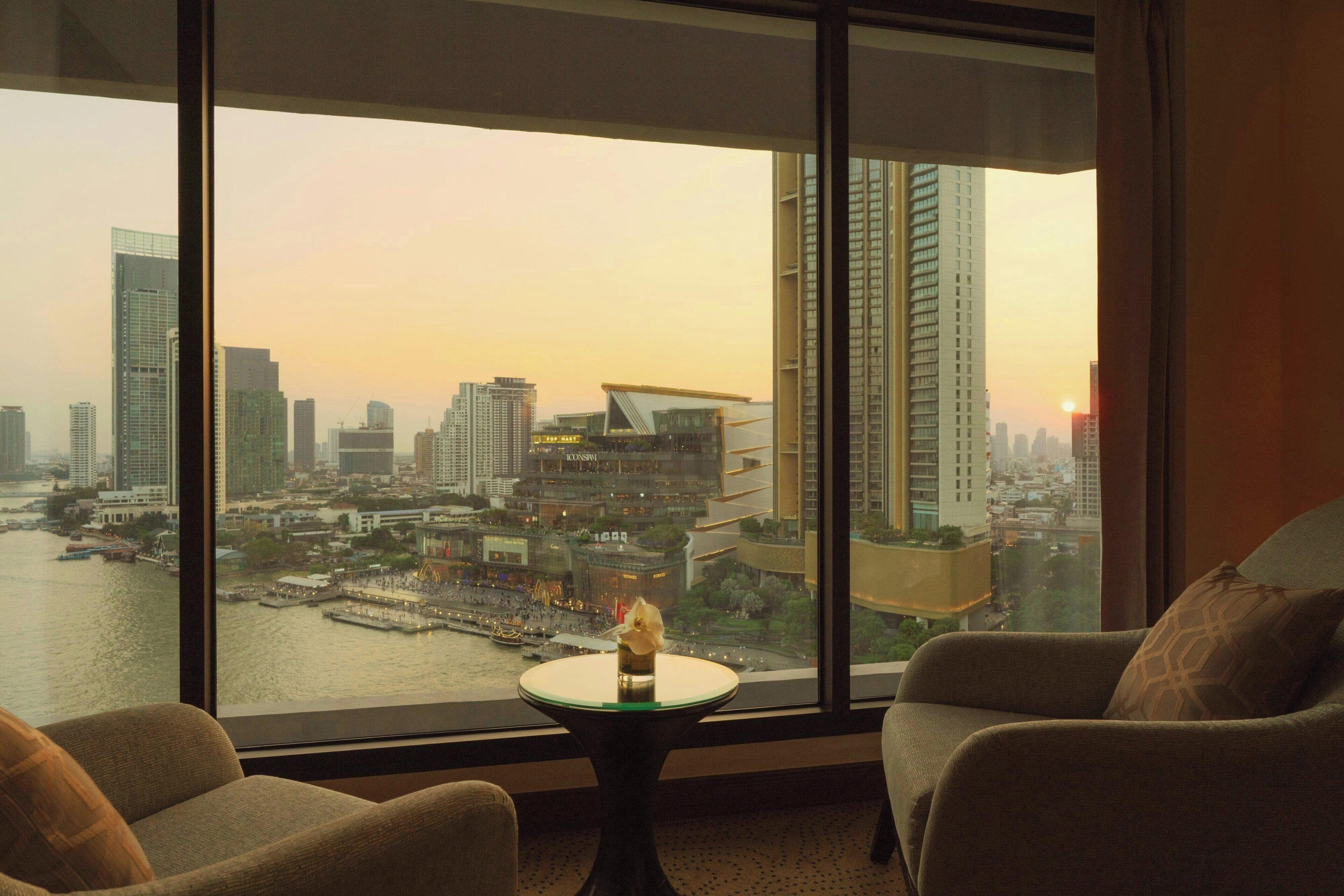 A room with a large window offering a view of a cityscape and a body of water at sunset. There is a small round table with a vase and a candle. Two armchairs are positioned in front of the window.