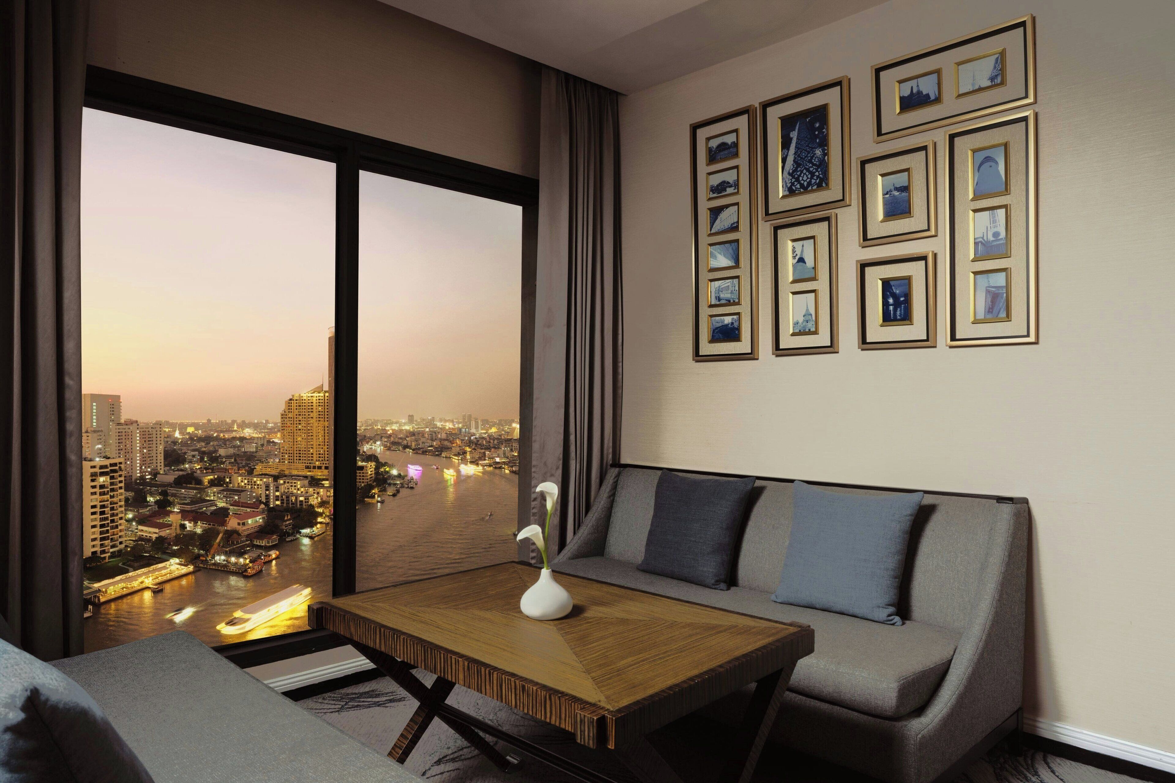 Habitación de hotel moderna con un ventanal amplio que ofrece vistas a la ciudad, un sofá, una mesita con un florero y cuadros en la pared.