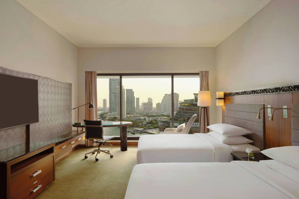 Habitación de hotel con dos camas, un escritorio, una silla, un televisor y un amplio ventanal con vista a la ciudad.