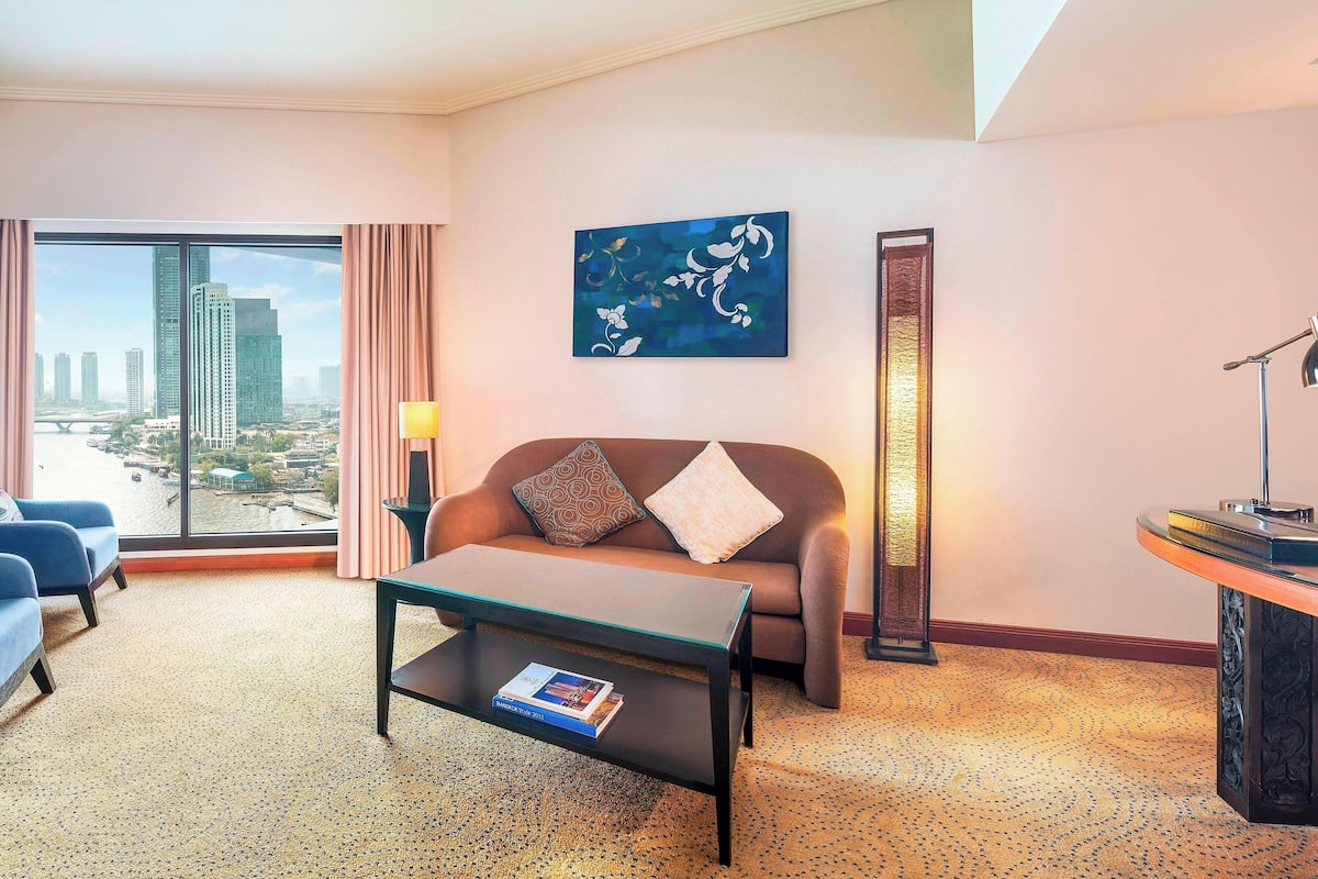 Habitación de hotel con un ventanal amplio que ofrece vistas a la ciudad, un sofá con cojines, una mesa de centro y una lámpara.