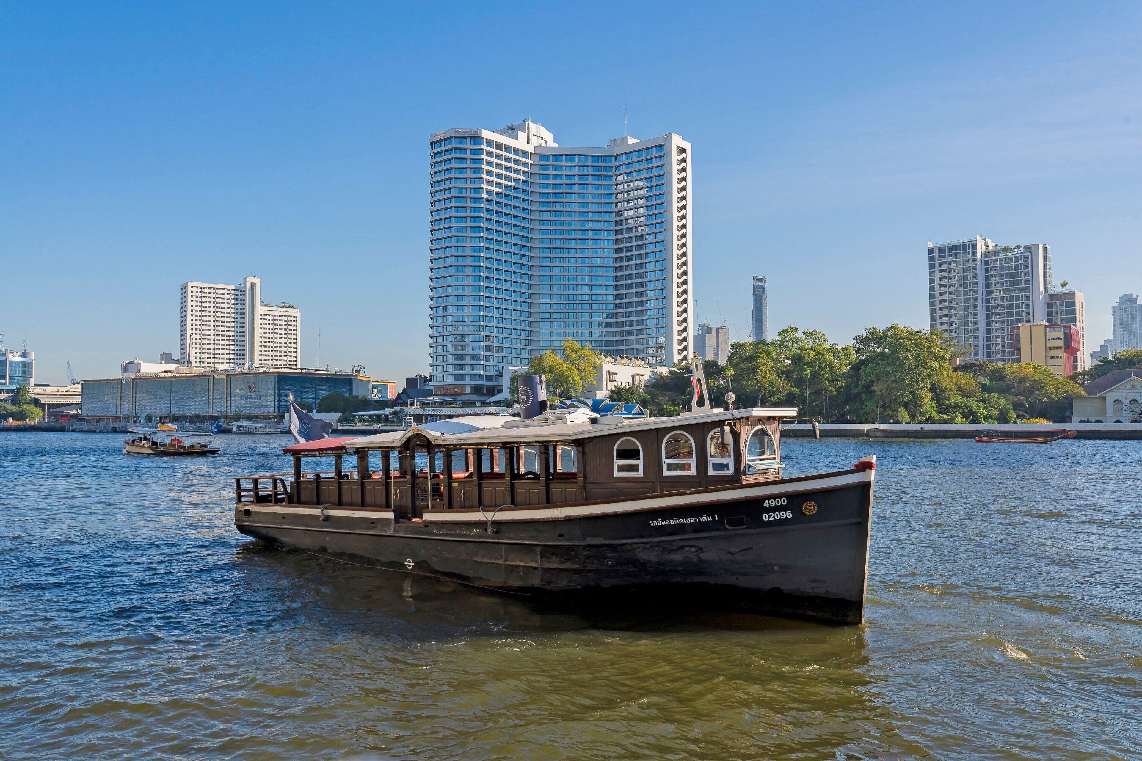 A riverboat with a cabin and deck seating, docked near a city skyline with high-rise buildings.