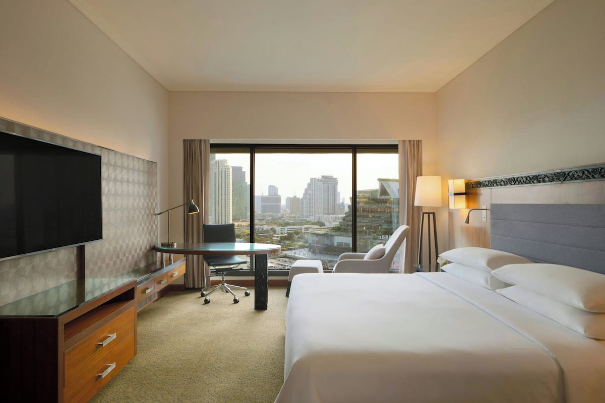 Habitación de hotel con cama, escritorio, silla, televisor y ventana grande con vista a la ciudad.