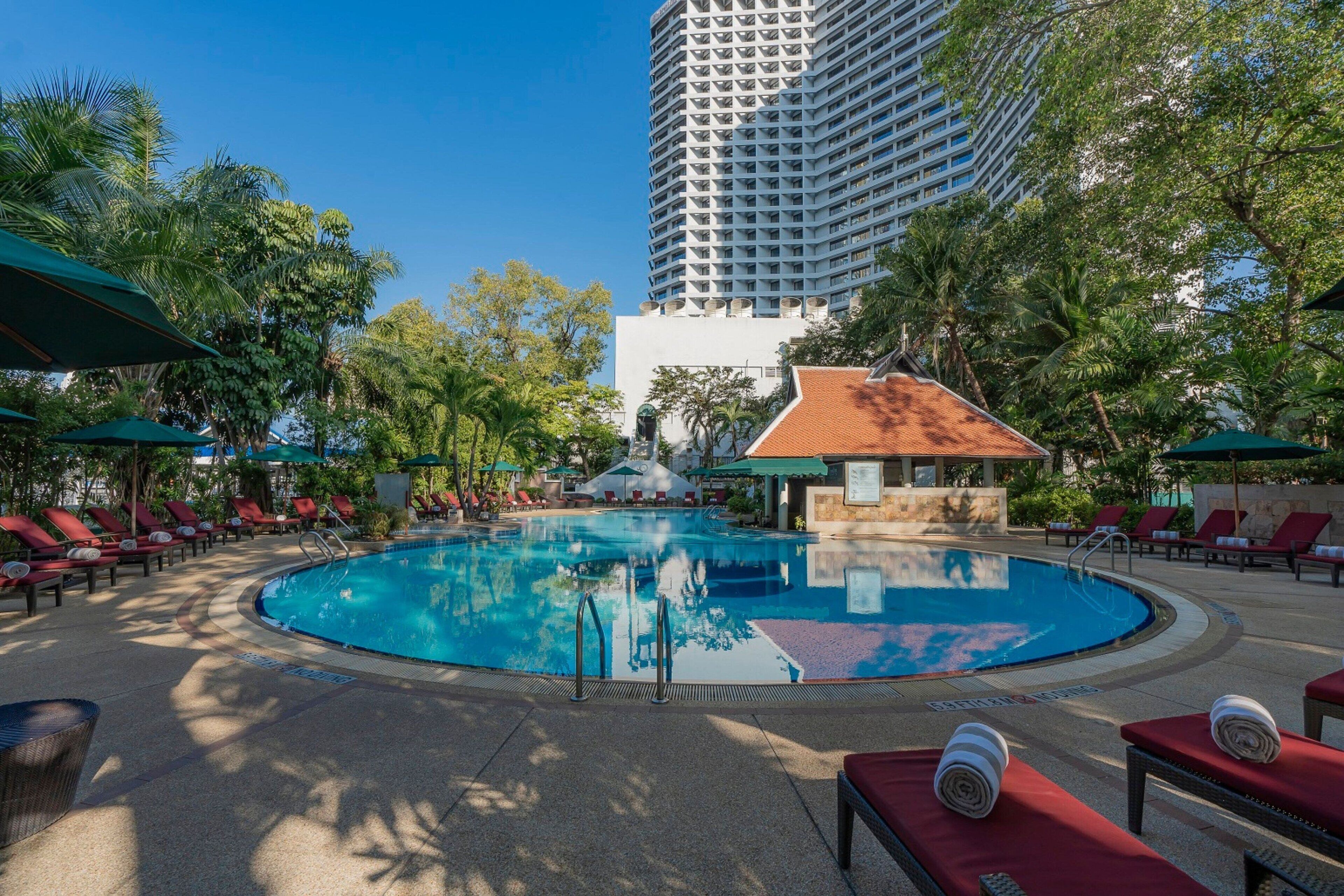Una piscina rodeada de tumbonas y sombrillas, con un edificio alto al fondo.