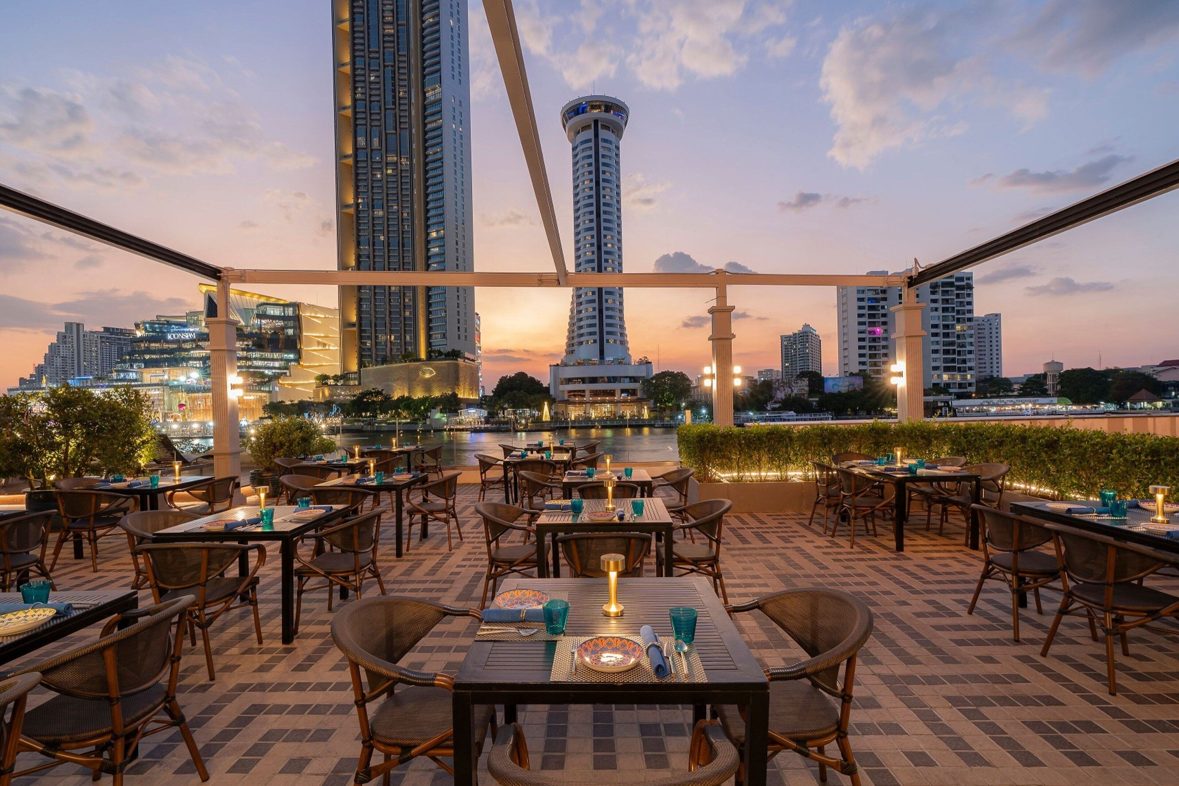 An outdoor dining area with tables and chairs, overlooking a cityscape at dusk.