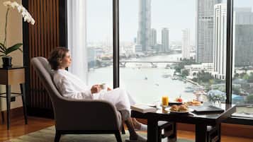 A person sitting in a chair by a window with a city view, a table with food and drinks, and a potted orchid.