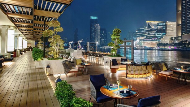 A rooftop bar with wooden flooring, seating arrangements, and a view of the city skyline and water at night. - Royal Orchid Sheraton Riverside Hotel Bangkok (Bangkok)