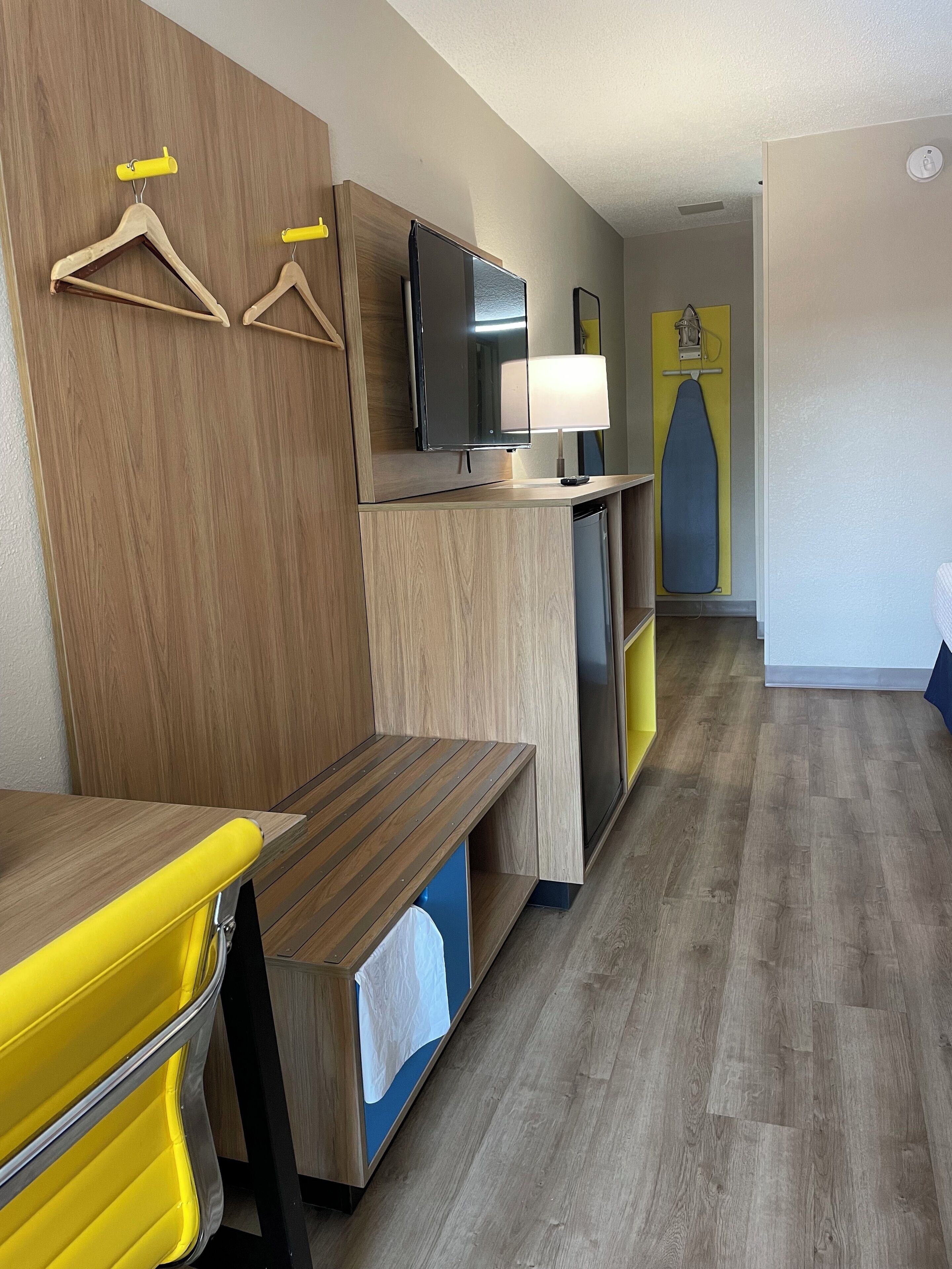 A modern hotel room with a wooden bench, a flat-screen TV, a lamp, and a yellow ironing board.